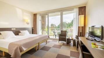 Ein Hotelzimmer mit Bett, Fernseher, Schreibtisch mit Stuhl und Balkon mit Blick auf Bäume.