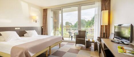 Habitación de hotel con cama, televisión, escritorio con silla y balcón con vistas a árboles.