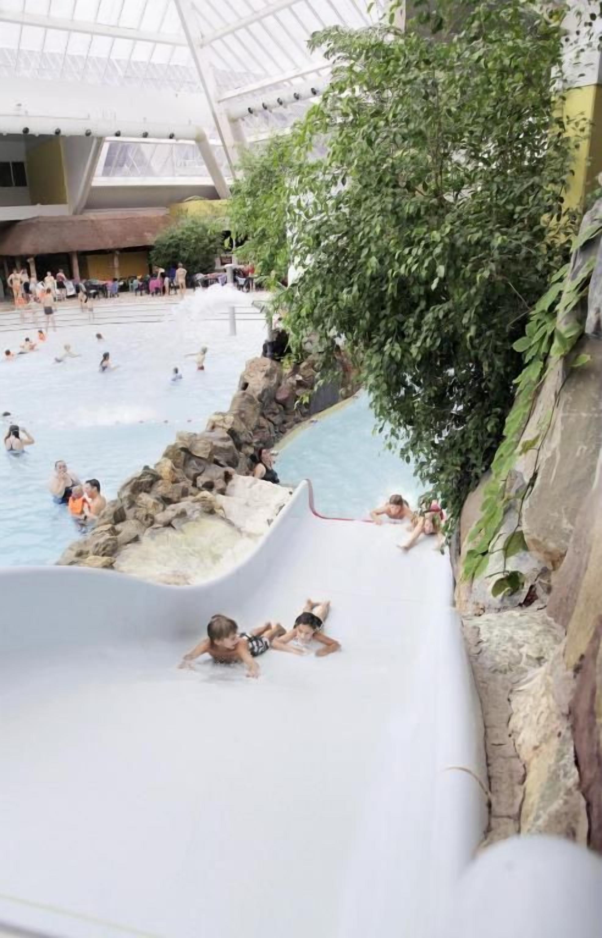 Un parque acuático interior con una piscina grande, toboganes y una cascada artificial de rocas.