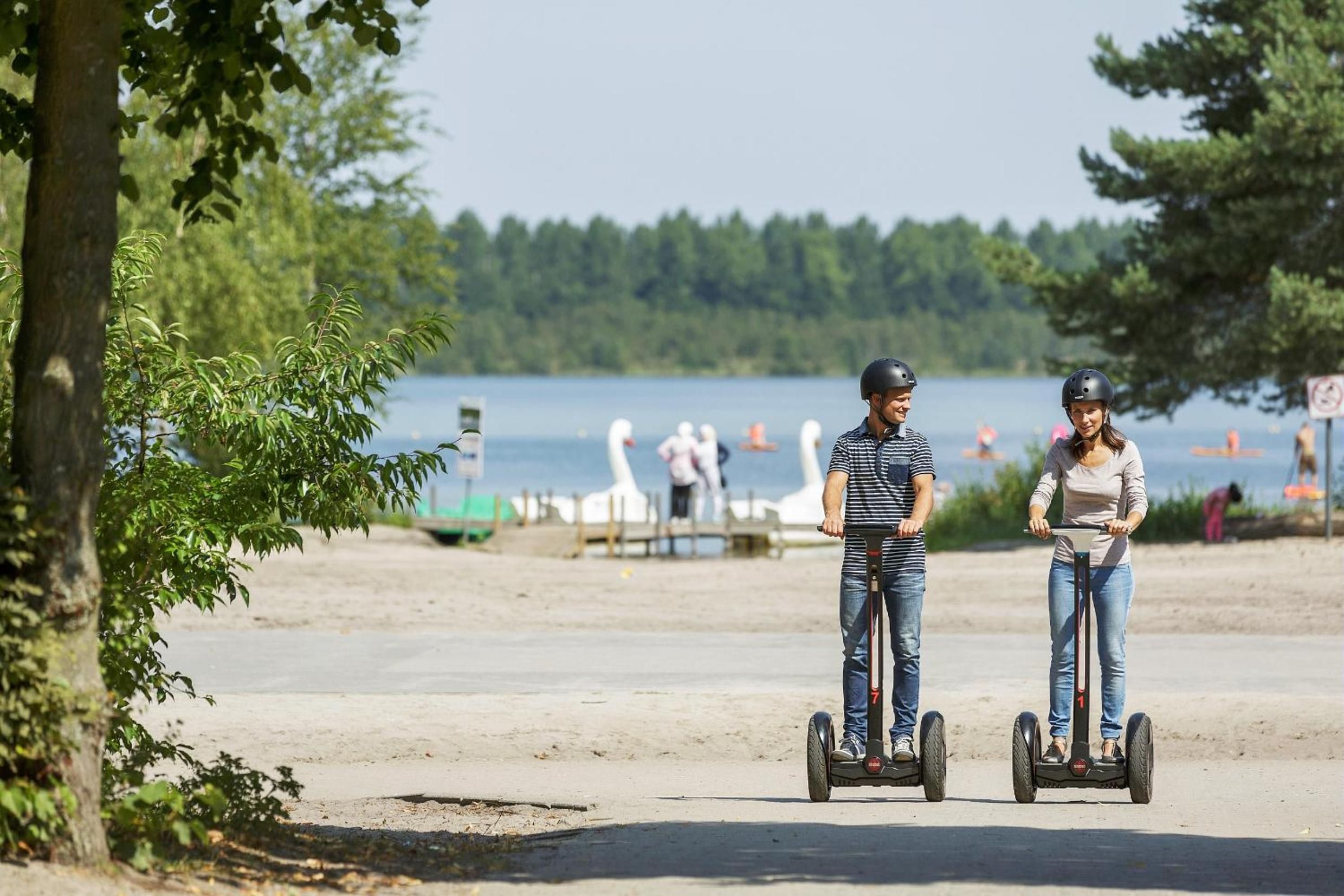 Dos personas en segways por un sendero arenoso, con un lago y árboles al fondo.