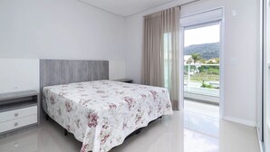 A bedroom with a bed, a nightstand, a dresser, and a sliding glass door leading to a balcony with a view of greenery and hills. - Costa Dourada 204 - Apto Praia Mariscal (Bombinhas)