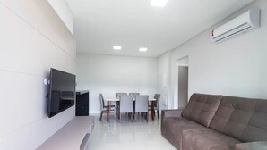 A modern living room with a flat-screen TV, a sofa, a dining table with chairs, and an air conditioning unit. - Costa Dourada 204 - Apto Praia Mariscal (Bombinhas)