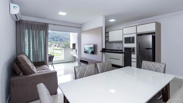 A modern kitchen with a flat-screen TV, stainless steel appliances, and a dining area with a white table and leather chairs. - Costa Dourada 204 - Apto Praia Mariscal (Bombinhas)