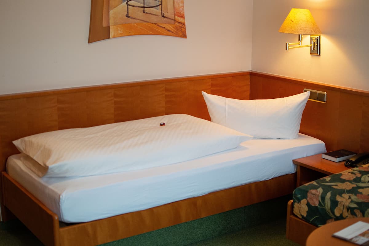 A neatly made bed with white linens, a wooden headboard, a nightstand with a lamp, and a small table with a book and a phone.