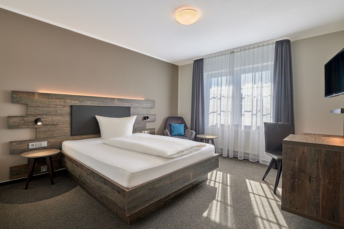 A modern hotel room with a large bed, a desk, a chair, and a TV.