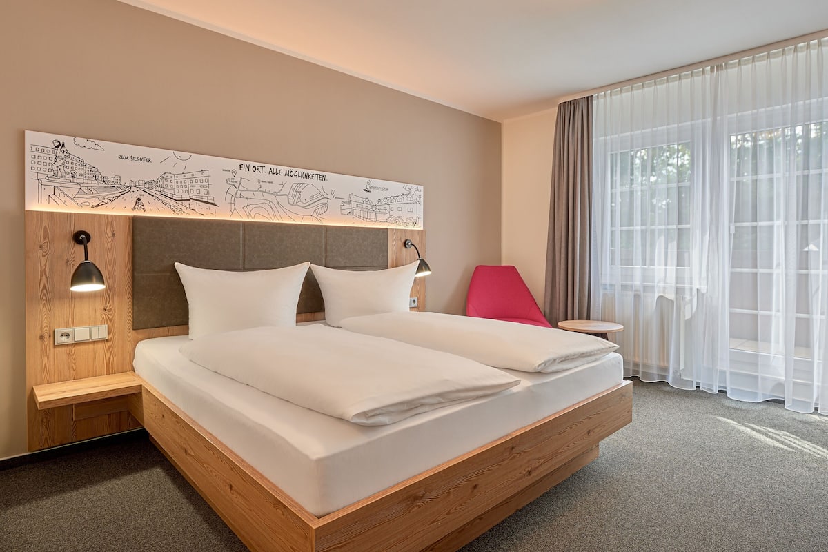 A hotel room with a large bed, a wooden headboard, a red chair, a small table, and a large window with sheer curtains.