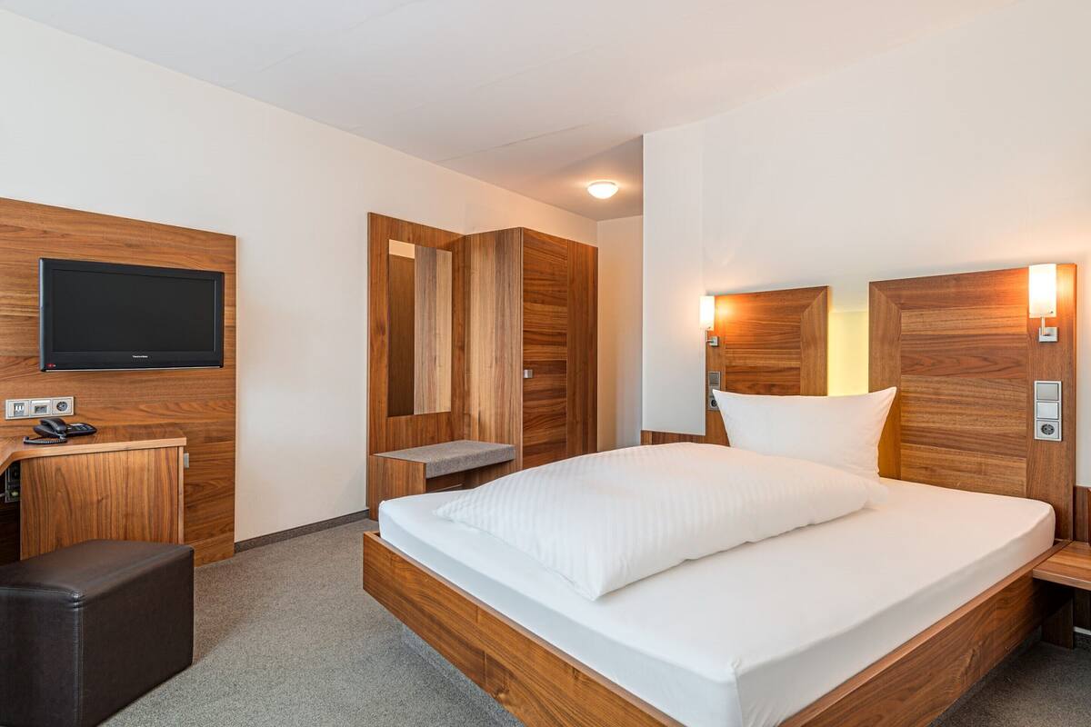 A modern hotel room with a large bed, wooden headboard, a flat-screen TV, a desk, and a small seating area.