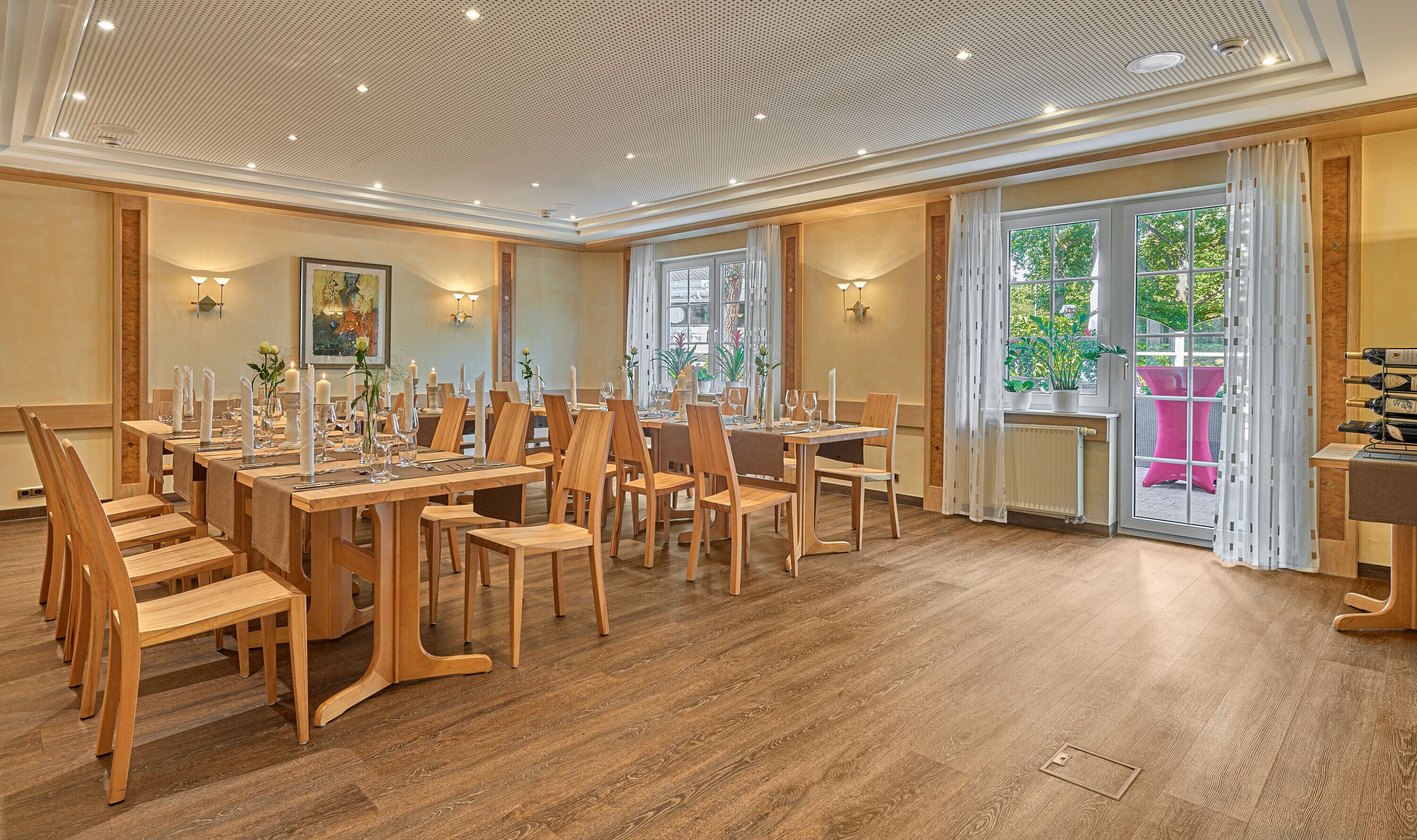 A dining area with wooden tables and chairs, a ceiling with recessed lighting, and a view of greenery outside.