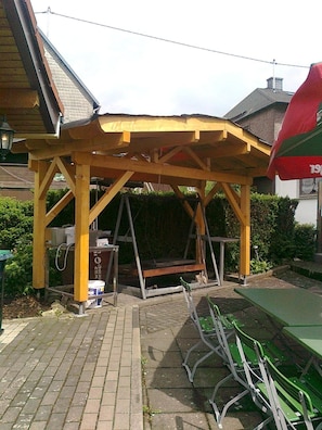 Una estructura de pérgola de madera con un columpio debajo, rodeada por un patio de ladrillo y sillas verdes.