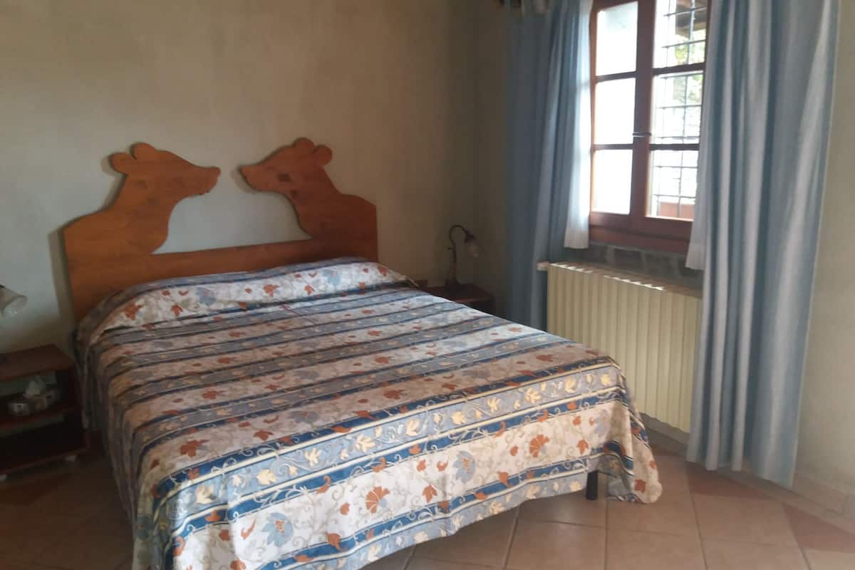 A bedroom with a wooden headboard, a patterned bedspread, a window with curtains, and a radiator.