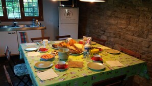 Une table à manger dressée avec différents plats et du pain, entourée de chaises ornées de coussins à motifs floraux, dans une pièce aux murs de pierre et dotée d’une fenêtre.