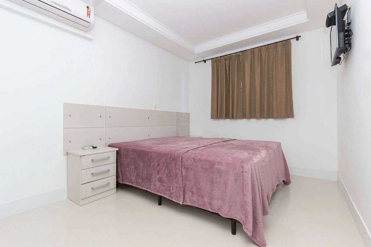 A bedroom with a bed, a nightstand, a dresser, a window with curtains, and an air conditioning unit.
