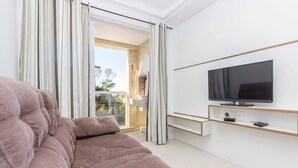 A modern living room with a large sofa, a flat-screen TV, and a sliding glass door leading to a balcony. - Madeira Aptos na Praia de Bombas (Bombinhas)