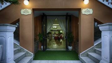 The entrance to a hotel with a grand staircase, a green carpet, and a well-lit interior visible through the glass doors.