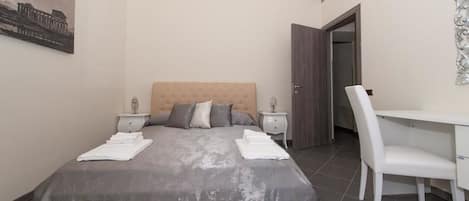 A bedroom with a large bed, a desk, and a chair. There is a door and a wall decoration.