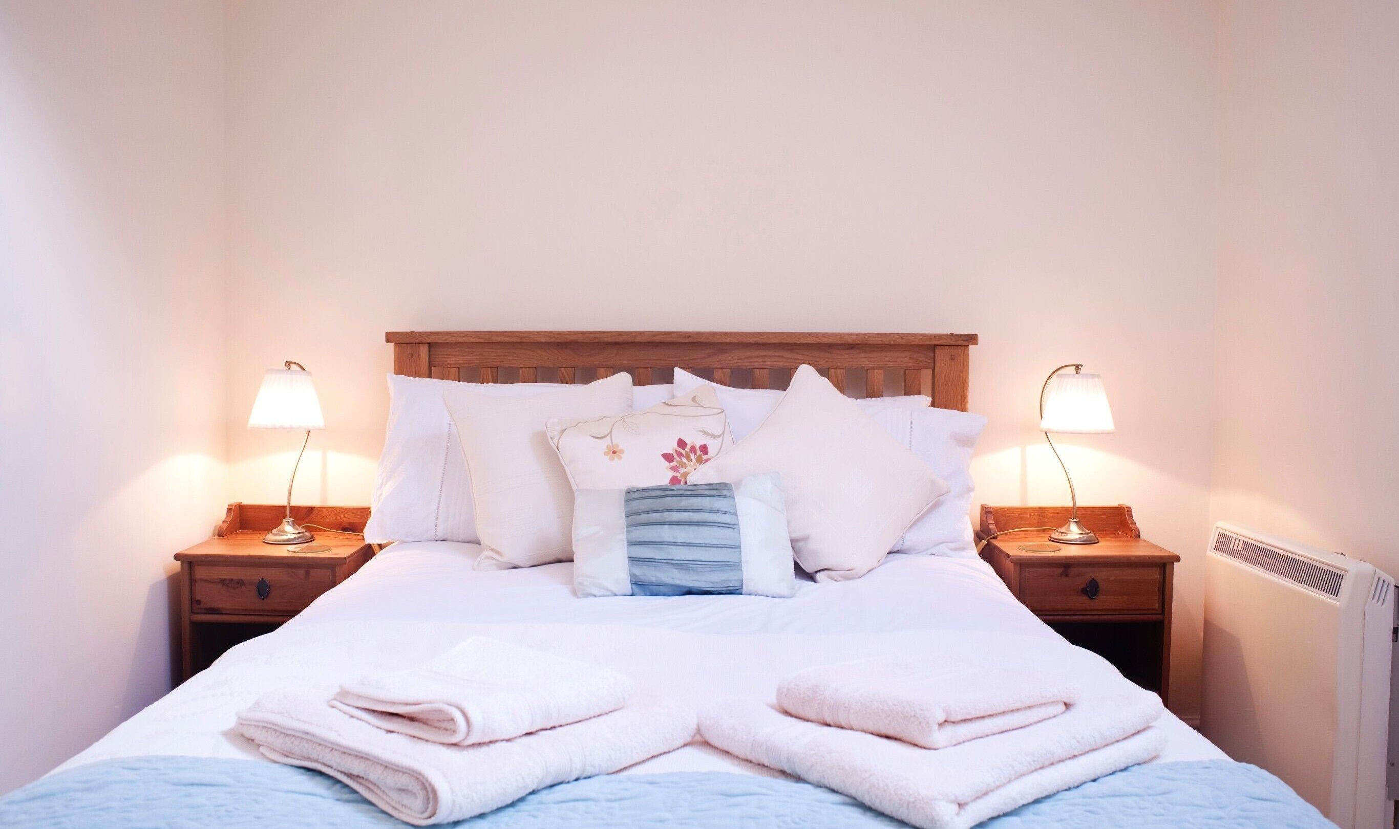 Una cama bien tendida con sábanas blancas y una manta azul, flanqueada por dos mesitas de noche con lámparas, un cabecero de madera y un radiador.