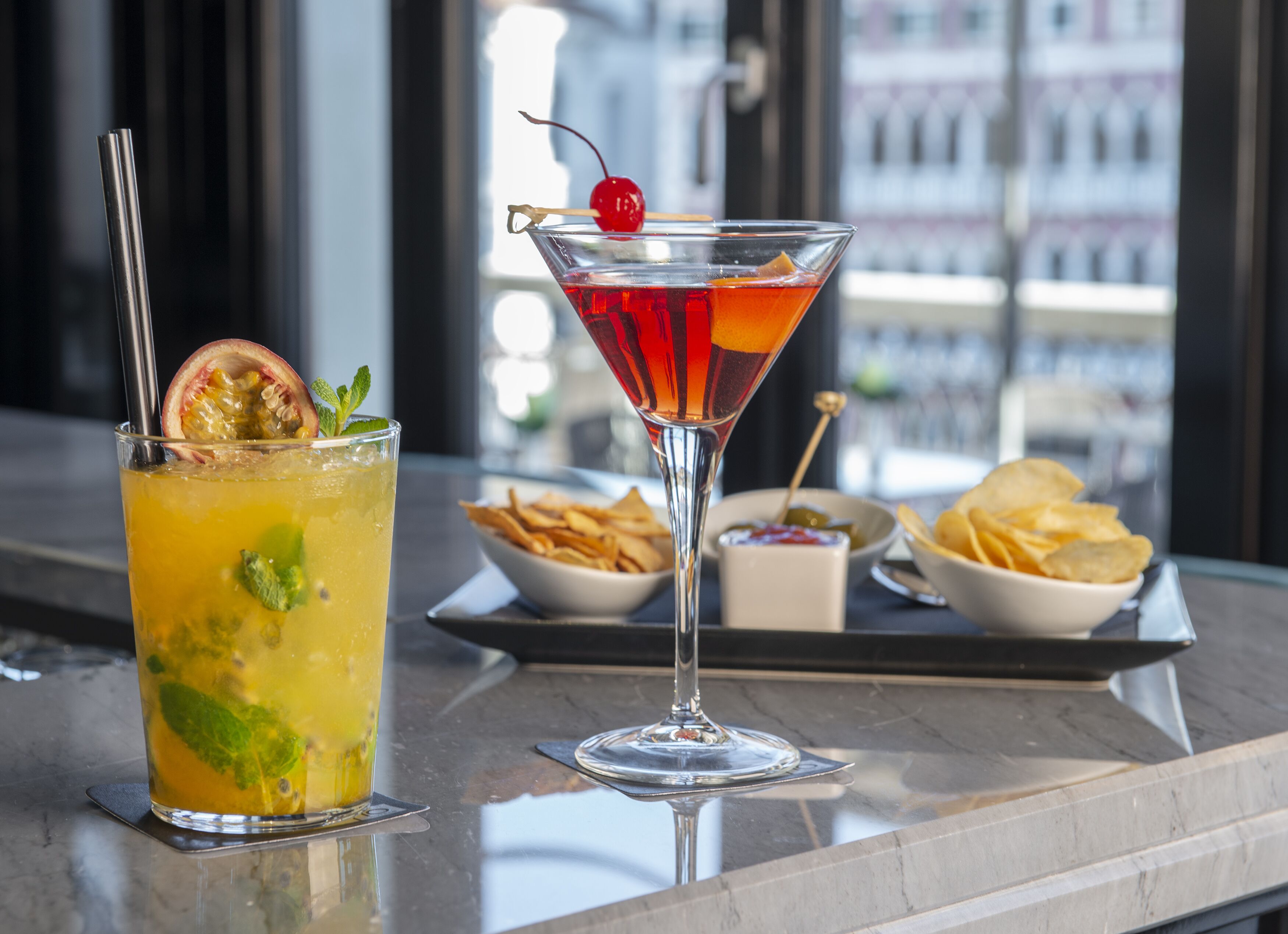 Two cocktails on a bar counter with a city view in the background.