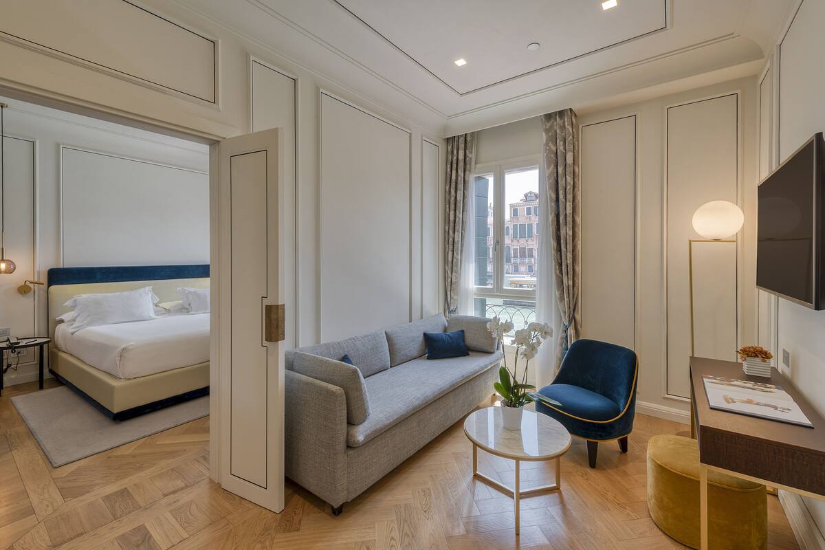 Habitación de hotel con una cama, un sofá gris, un sillón azul, una mesita redonda y un televisor.