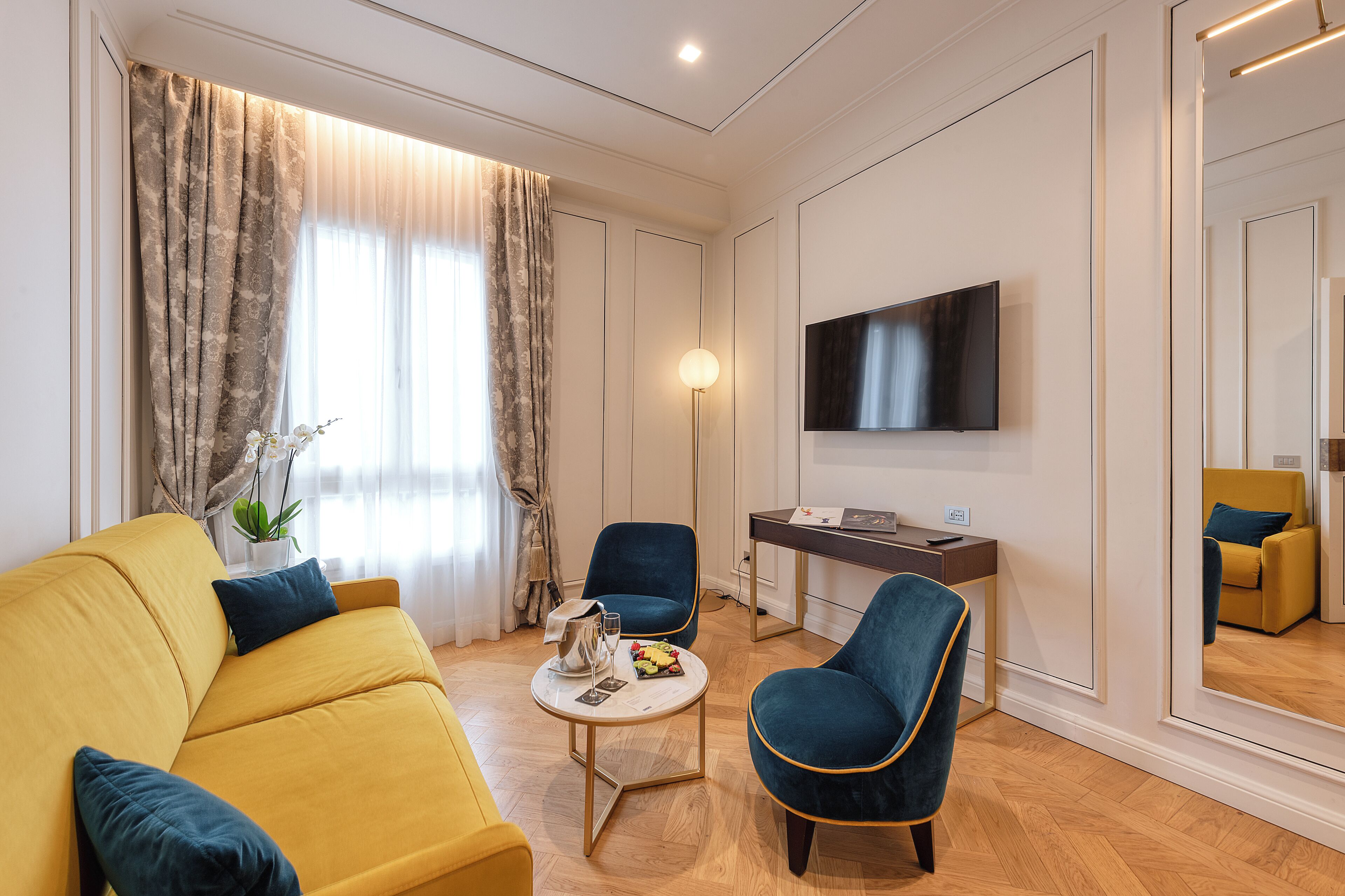 A modern living room with a yellow sofa, a blue armchair, a small round table with a lamp, and a flat-screen TV mounted on the wall.