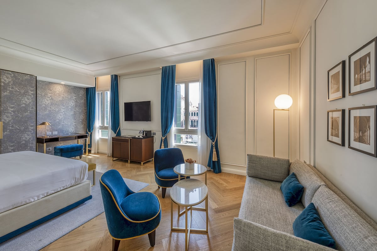 Una habitación de hotel con cama, sofá, dos sillones, una mesita, escritorio, televisión y ventana con cortinas.
