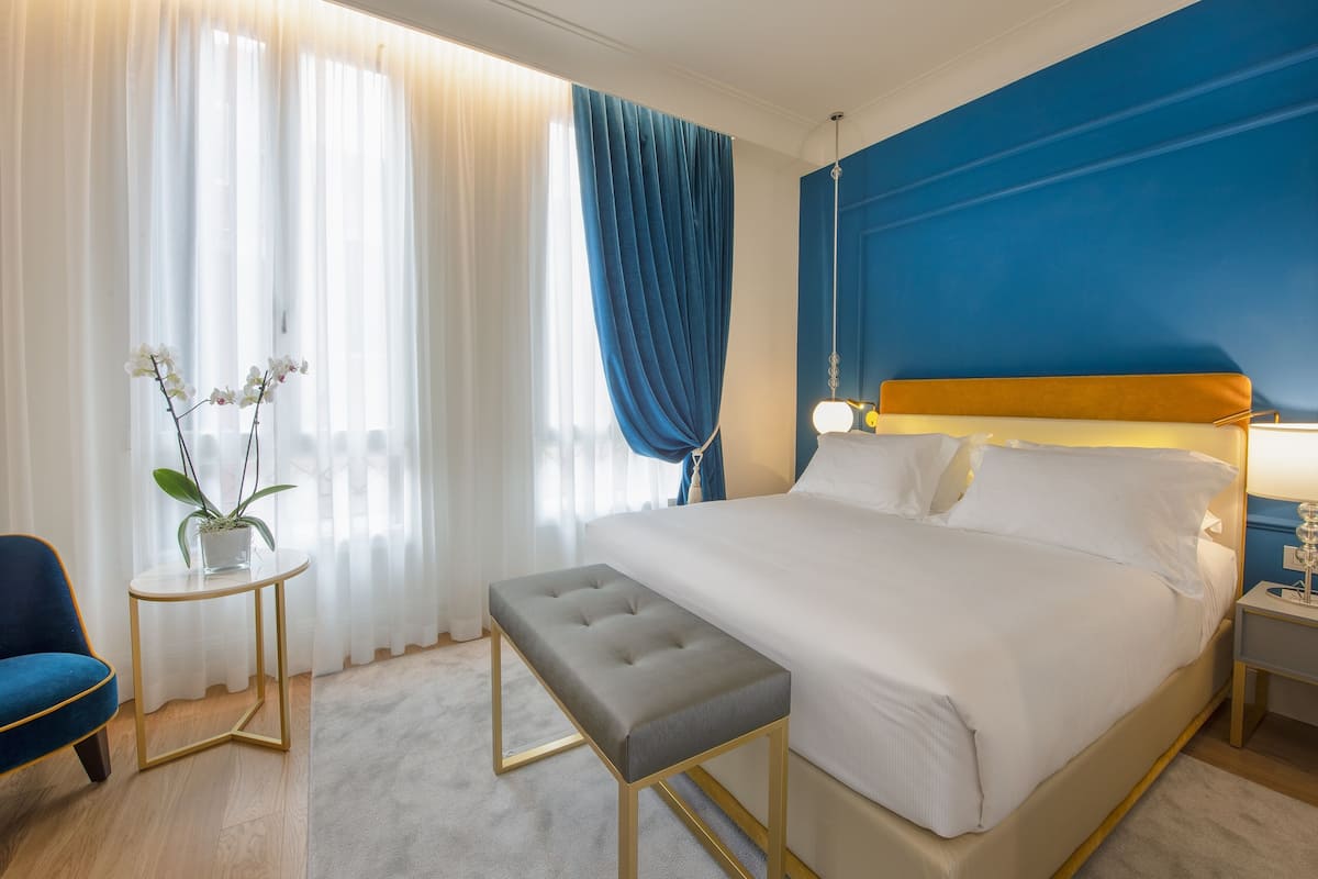 Habitación de hotel con cama, mesita de noche, banco, silla y planta en maceta.