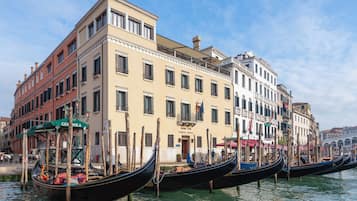 Une rangée de gondoles est amarrée le long d’un canal à Venise, avec des bâtiments historiques et un ciel dégagé en arrière-plan.