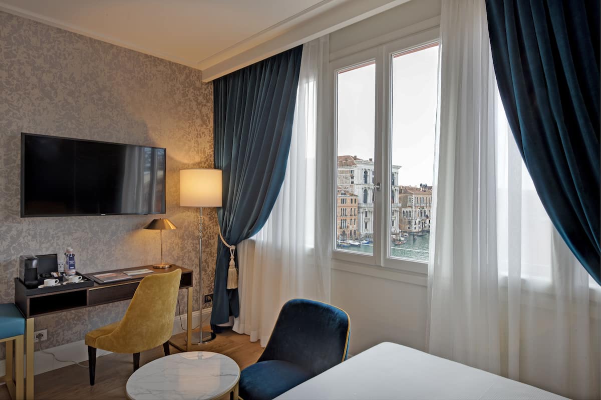 Habitación de hotel con una cama, un escritorio con televisor de pantalla plana, una silla, una lámpara y vista a un paisaje urbano por la ventana.