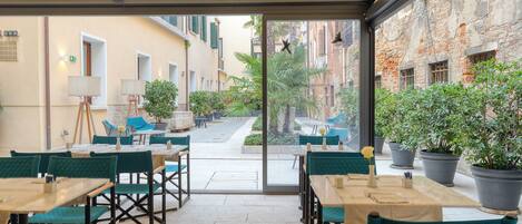 An outdoor café with tables and chairs, potted plants, and a view of the street.
