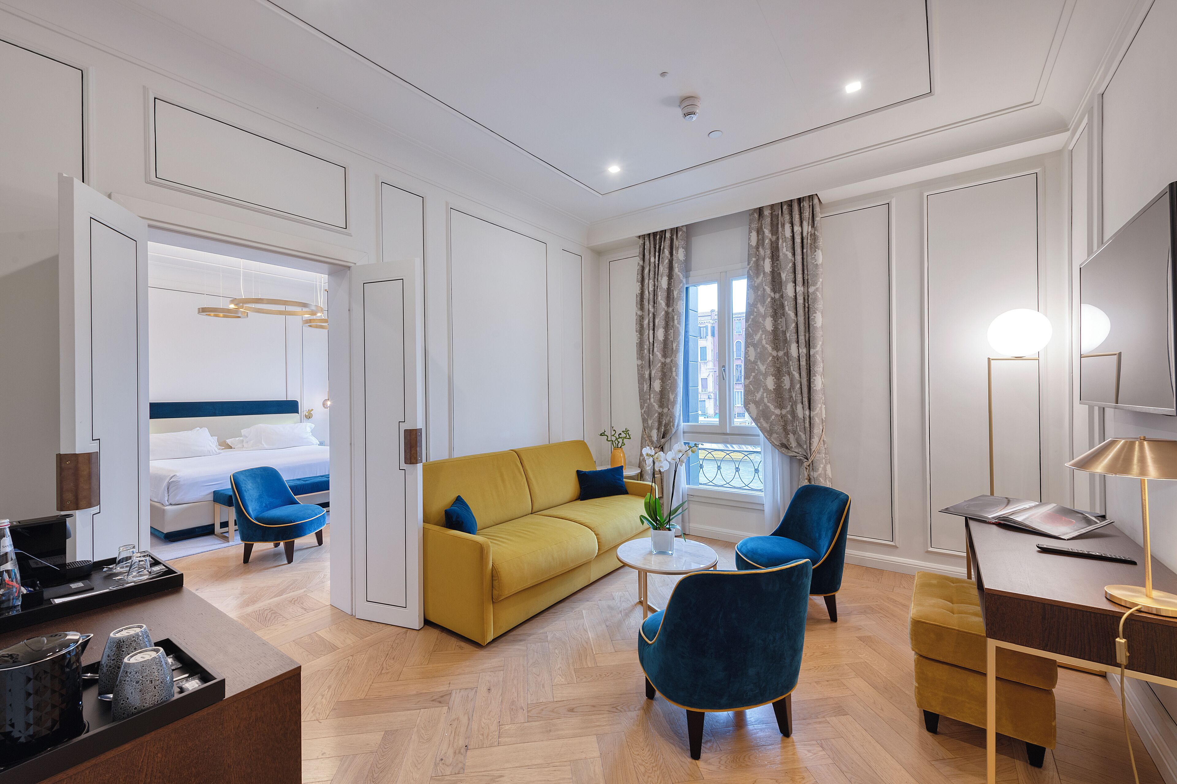 A modern hotel room with a yellow sofa, blue armchairs, a small round table, and a desk with a lamp.