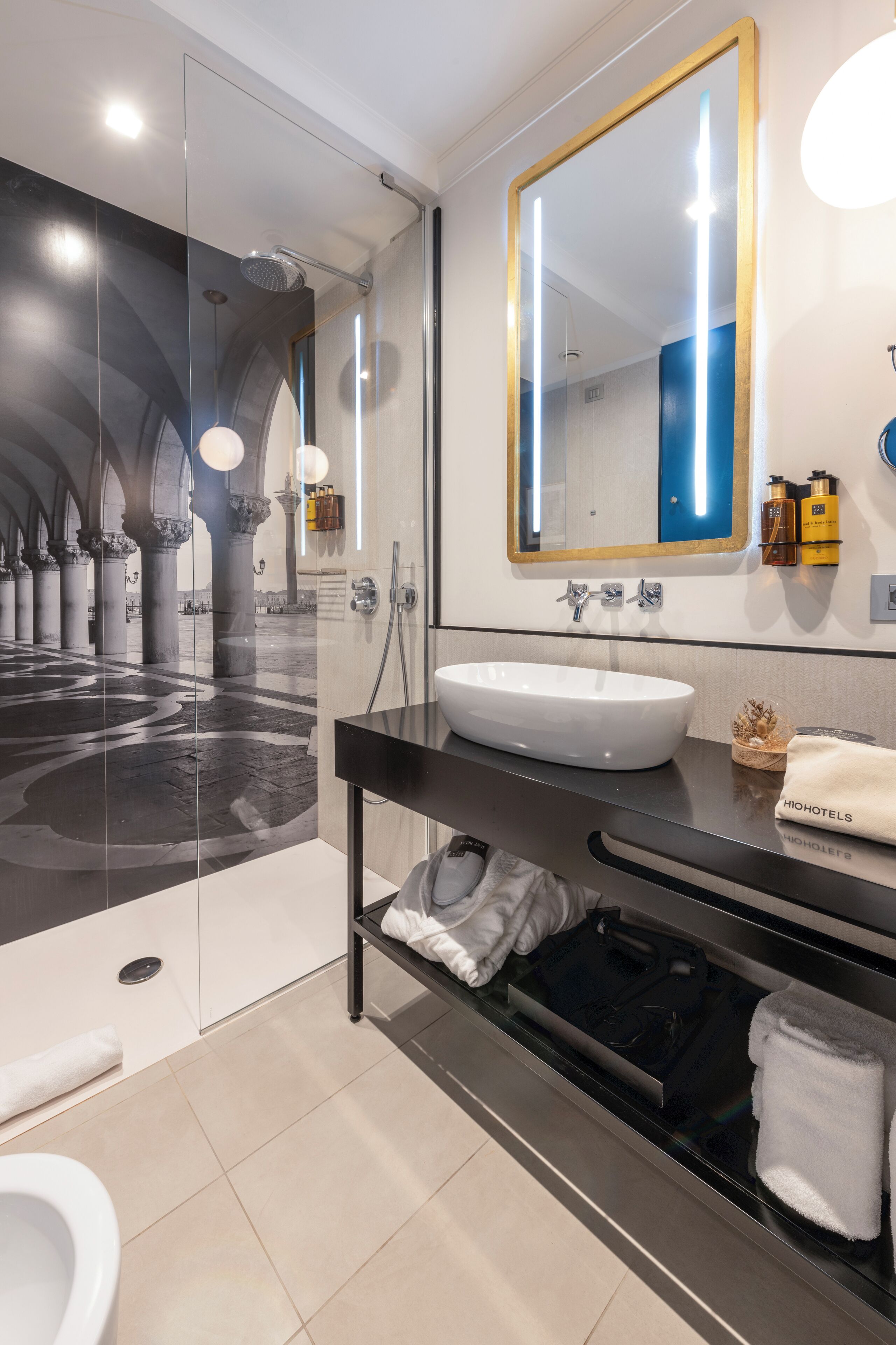 Ein modernes Badezimmer mit einer Glaskabine zur Dusche, einem weißen Waschbecken und einem großen Spiegel mit goldener Rahmung.