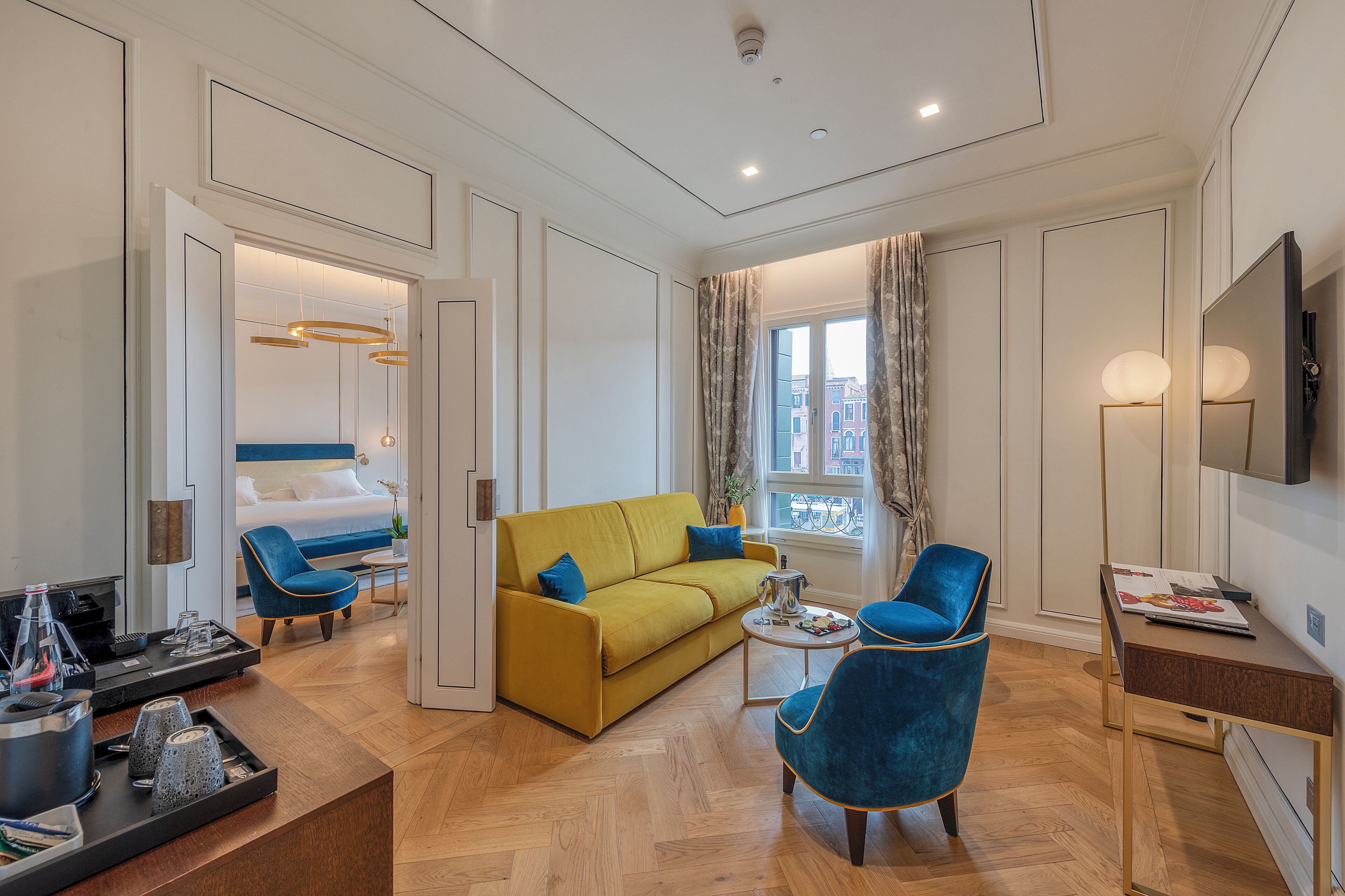 A modern hotel room with a yellow sofa, blue armchairs, a wooden coffee table, and a TV on a wooden stand.