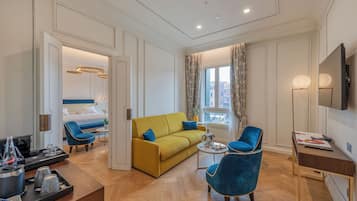 A modern hotel room with a yellow sofa, blue armchairs, a wooden coffee table, and a TV on a wooden stand.