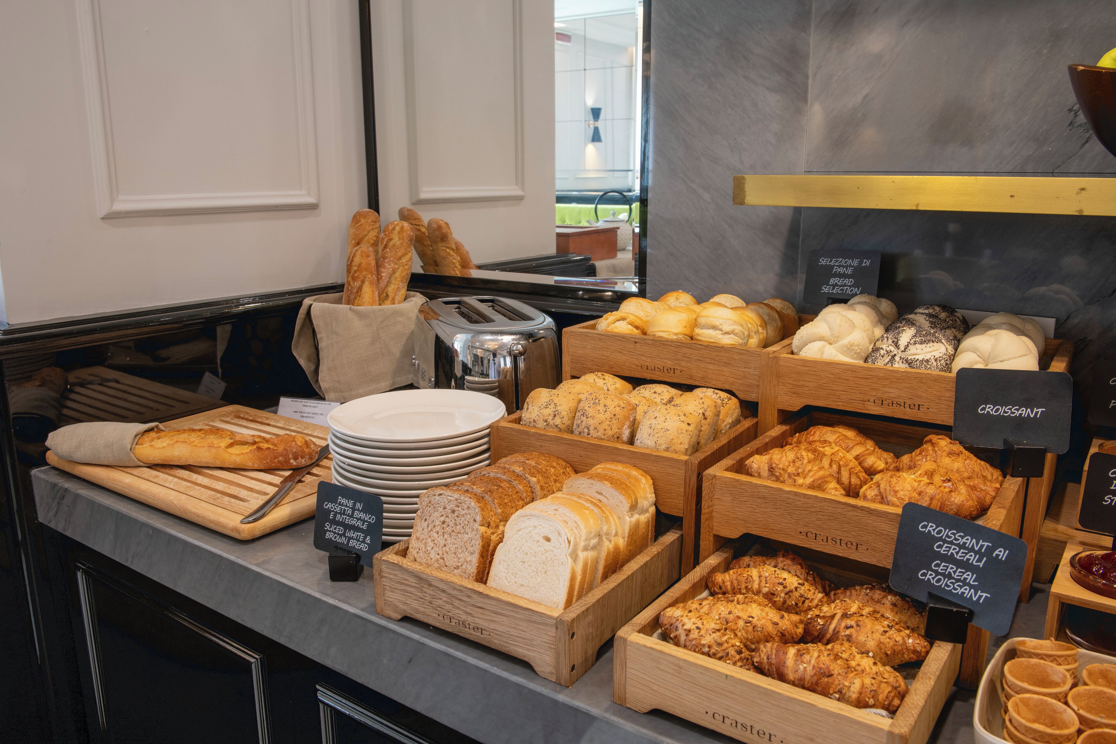 Mostrador de panadería con diversos panes y pasteles en cajas de madera, una tostadora y un cartel que indica la selección de pan.
