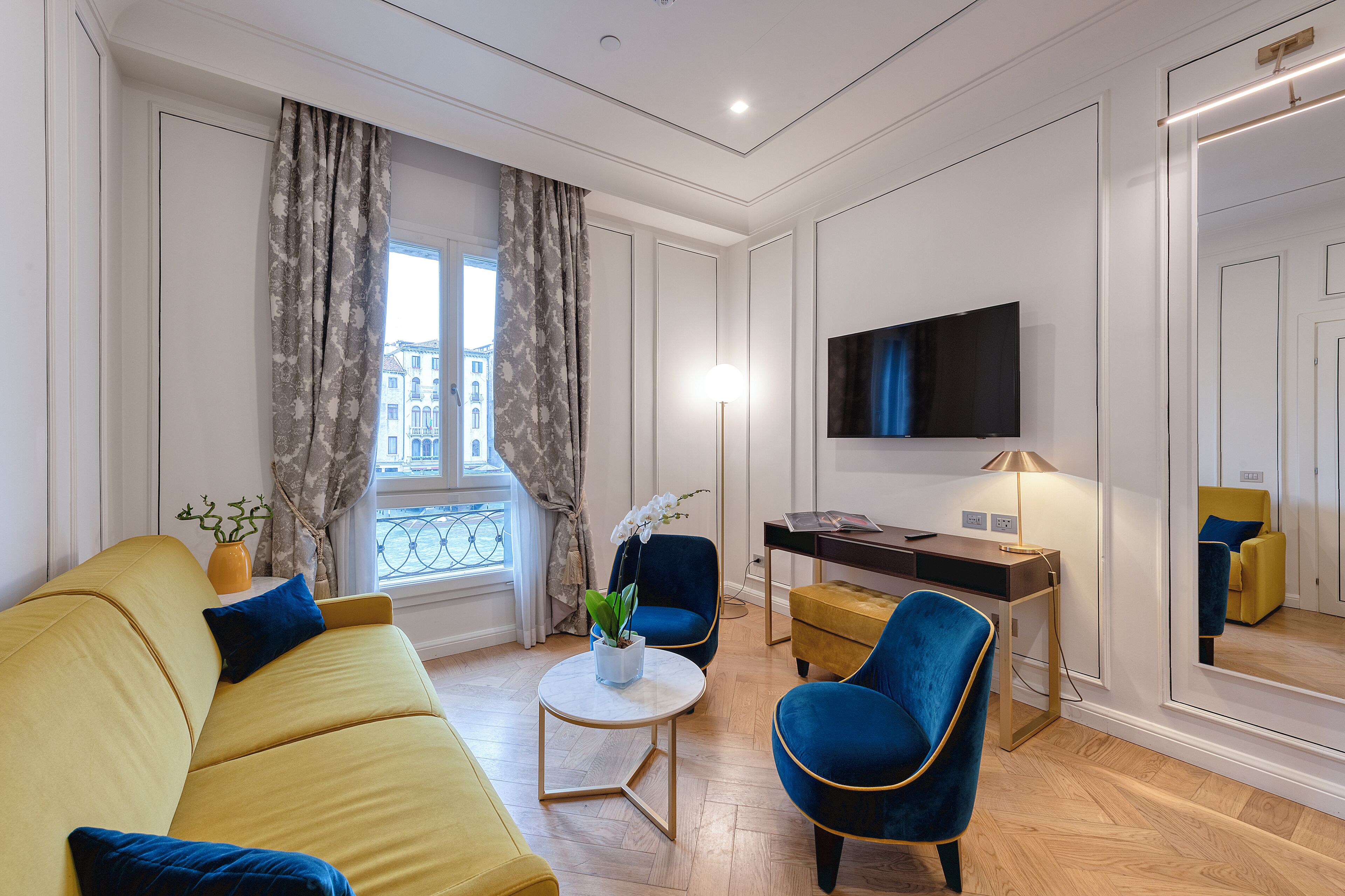 Una sala de estar moderna con un sofá amarillo, un sillón azul, una mesa redonda de centro y un escritorio con una lámpara.