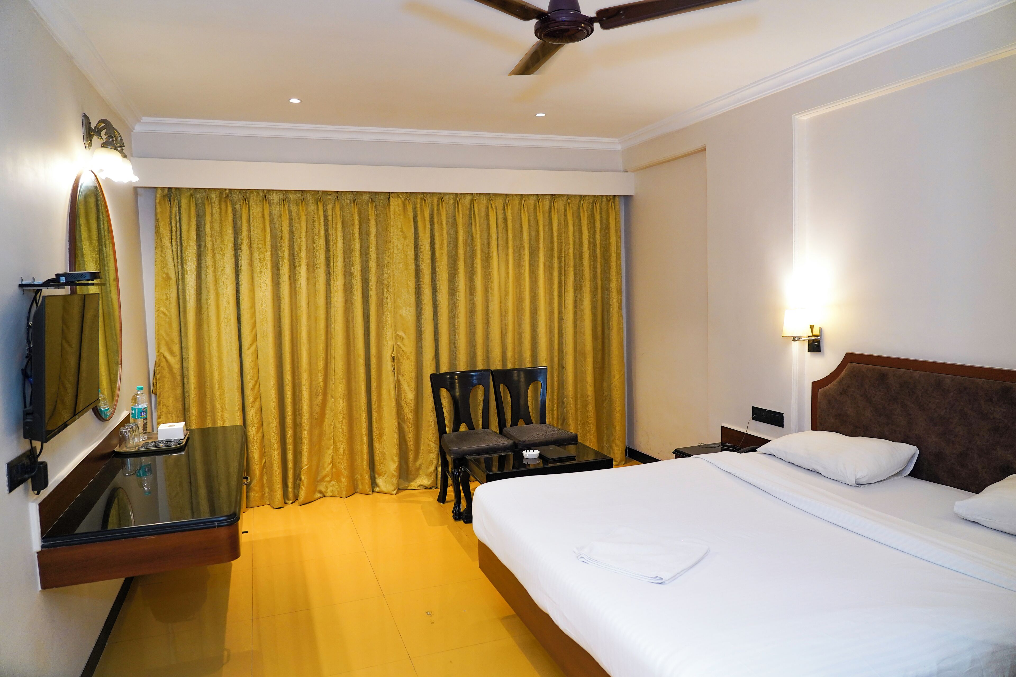 A hotel room with a bed, a desk with a chair, a mirror, and a ceiling fan.