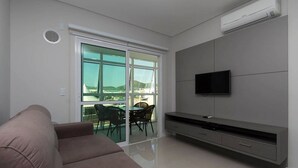 Un salon moderne comprenant un canapé, une table à manger avec des chaises, un téléviseur à écran plat fixé au mur et une porte-fenêtre coulissante donnant sur un balcon.