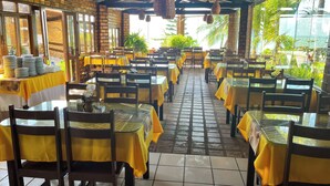 Un restaurante con varias mesas y sillas, cubierto por un techo, y amplias ventanas que dejan entrar luz natural.