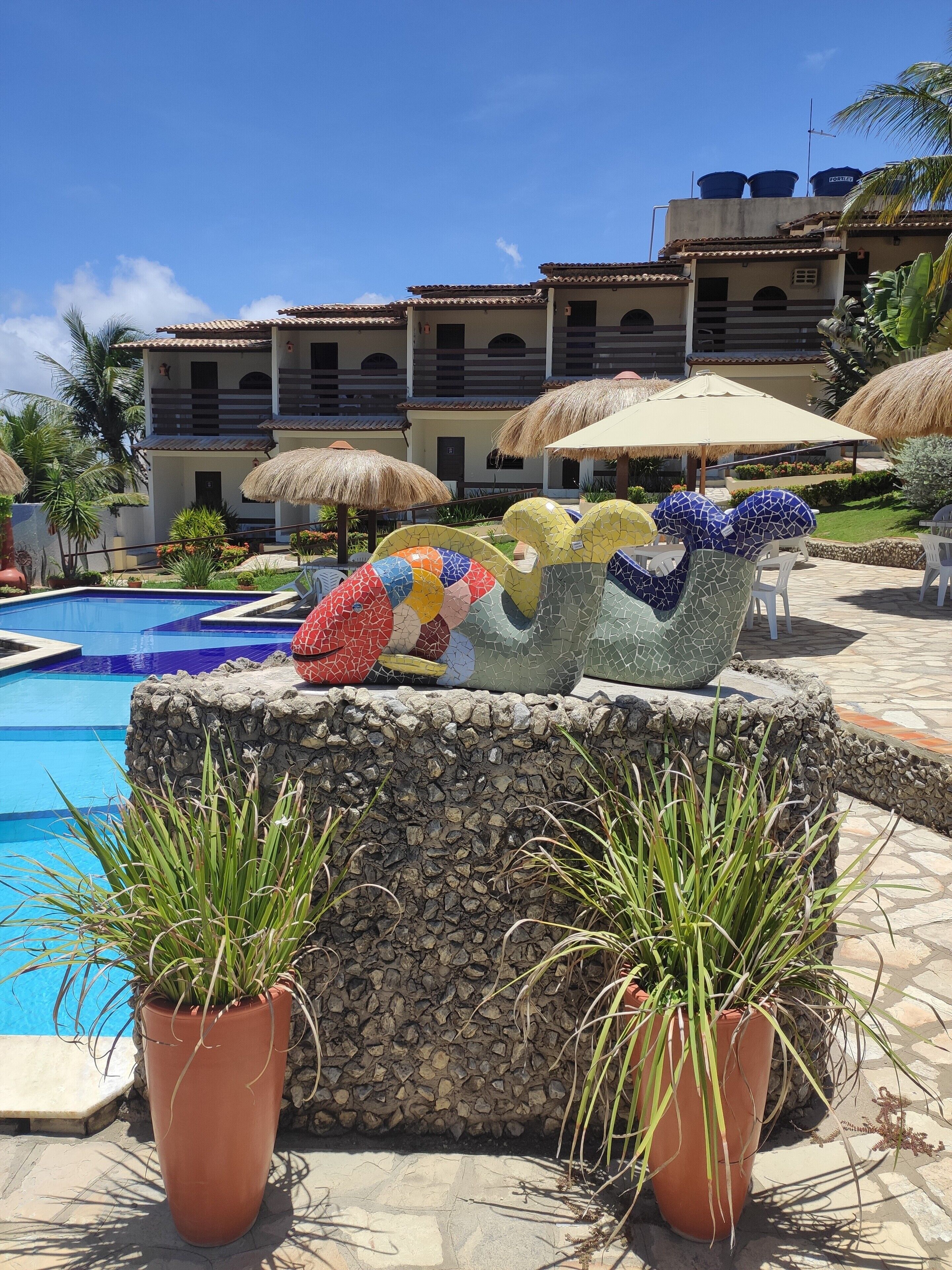 Uma piscina com parede de pedra e plantas em vasos, cercada por um edifício com telhado de azulejos e guarda-sóis de palha.