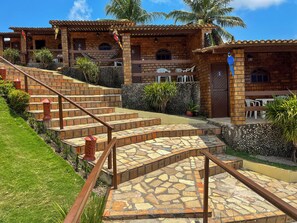 A multi-story building with a brick facade, outdoor seating area, and a stone staircase leading to the entrance. - Pousada Enseada do Sol (Conde)