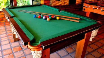 A pool table with balls and cues, surrounded by a brick wall and a wooden table with a rope barrier.