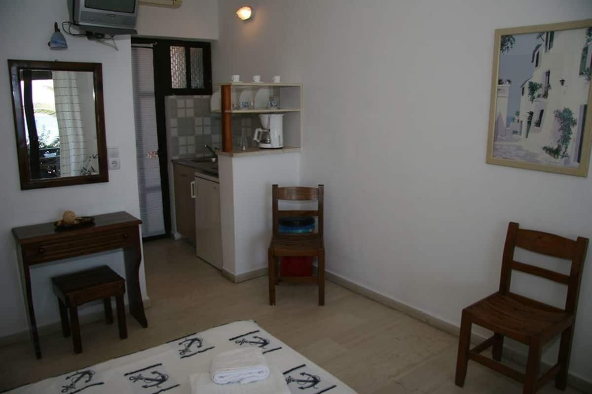 A compact room with a kitchenette, a wooden chair, a small table, and a painting on the wall.