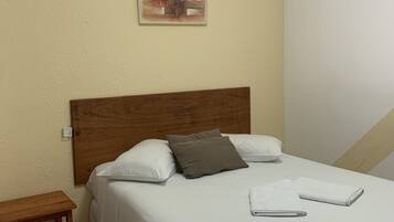 Ein ordentlich bezogenes Bett mit weißer Bettwäsche und zwei Kissen, ein Nachttisch daneben sowie ein kleines Bild an der Wand.