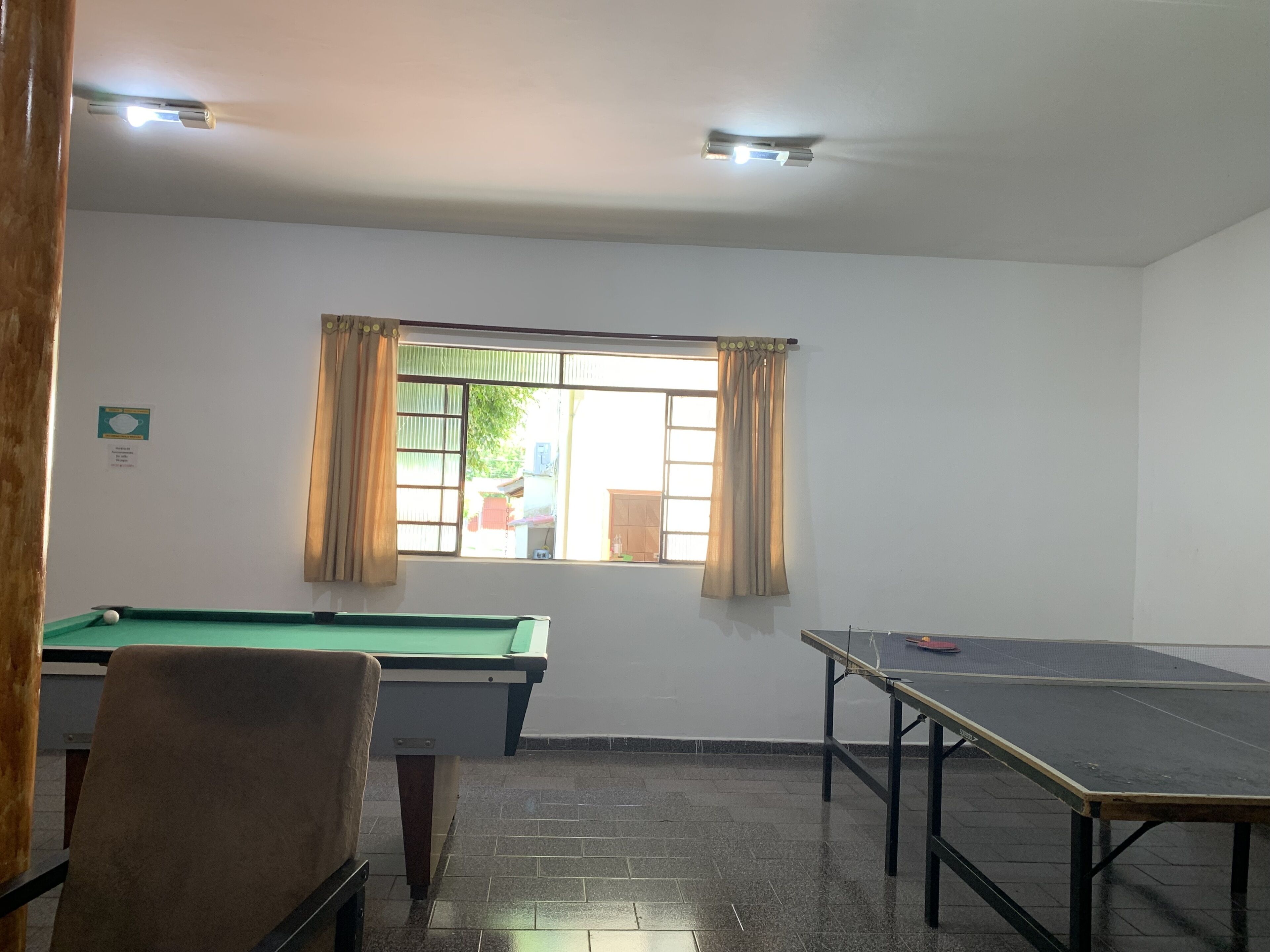 A room with a pool table, a billiards table, a green pool table, and a window with curtains.