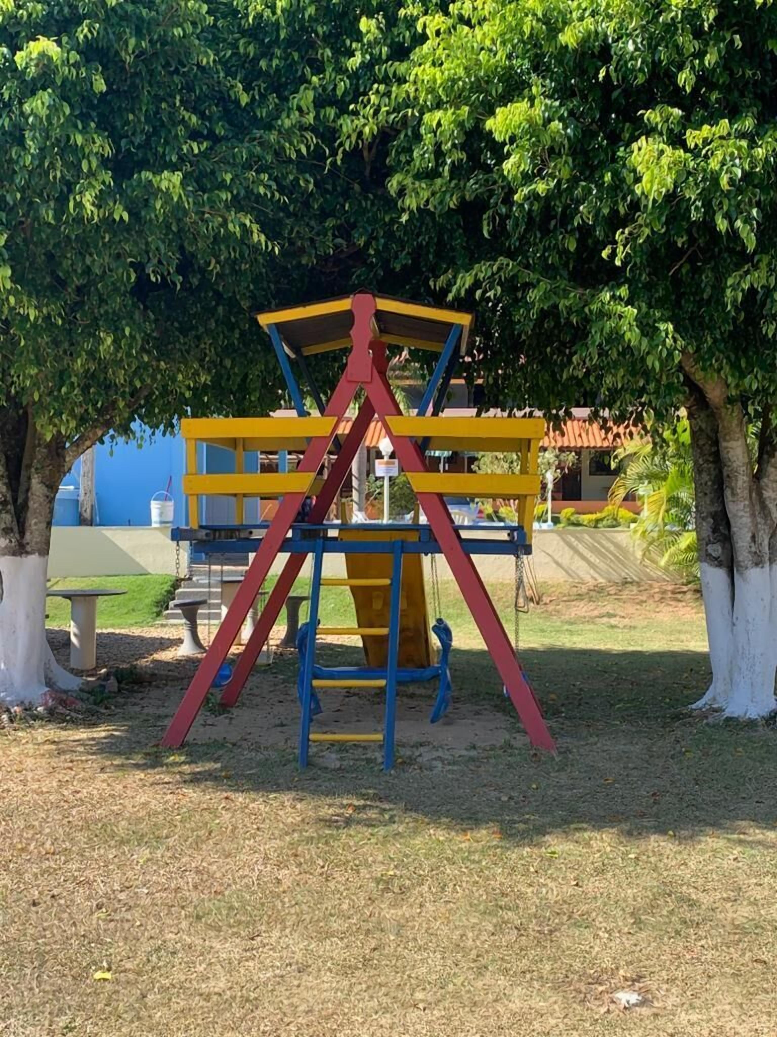 Un parque infantil con una estructura de escalada amarilla y roja, rodeado de árboles y un edificio azul al fondo.