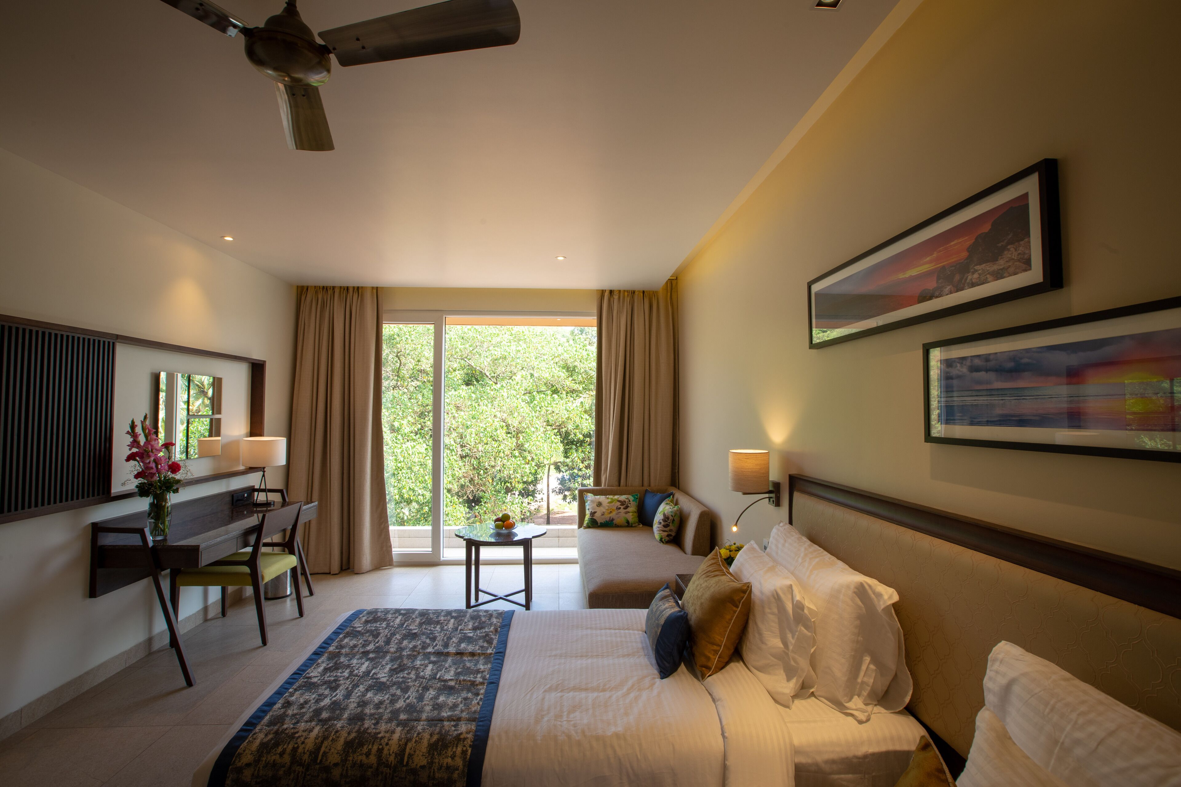 A hotel room with a bed, desk, chair, and a view of greenery outside.
