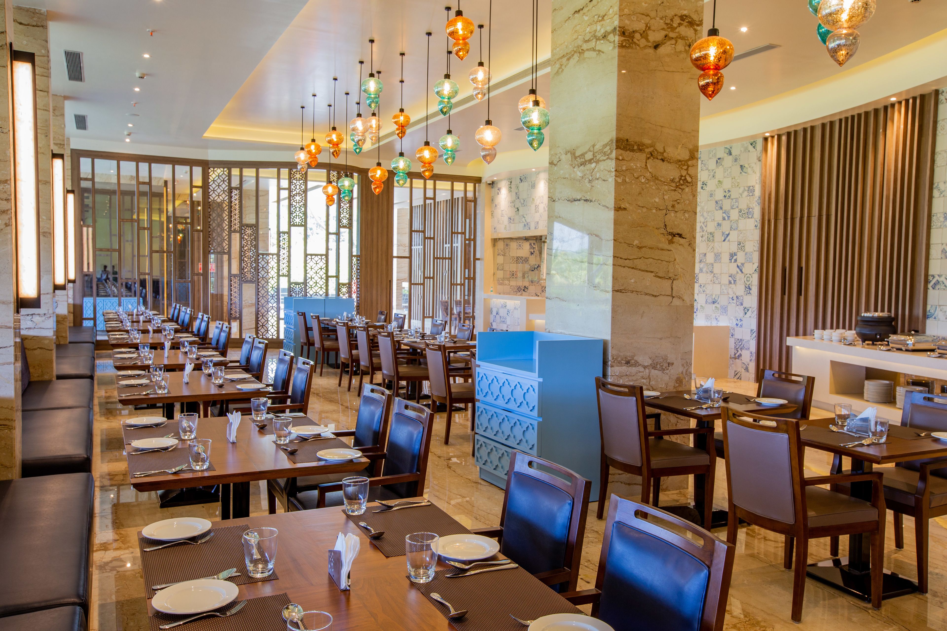 Un comedor amplio con varias mesas y sillas, lámparas decorativas colgantes y vistas al área de la cocina.
