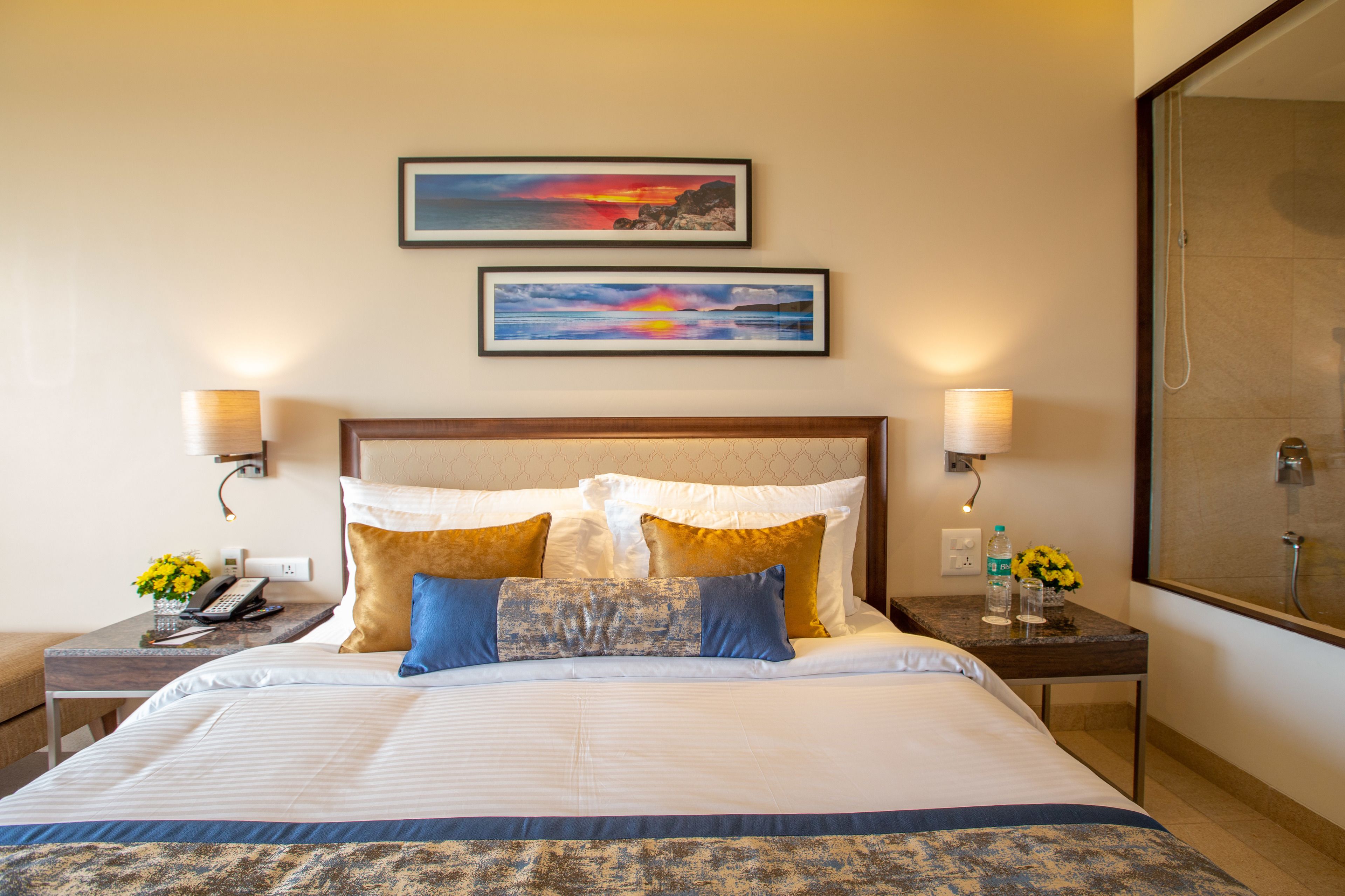 A hotel room with a large bed, two wall-mounted lamps, a nightstand with a phone and a vase of flowers, and two framed pictures on the wall.