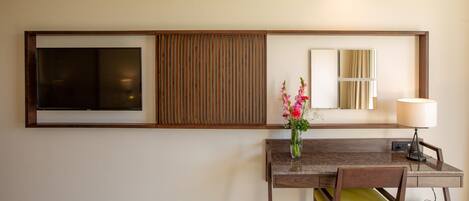A hotel room with a wooden desk, a chair, a vase with flowers, a lamp, and a TV mounted on the wall.