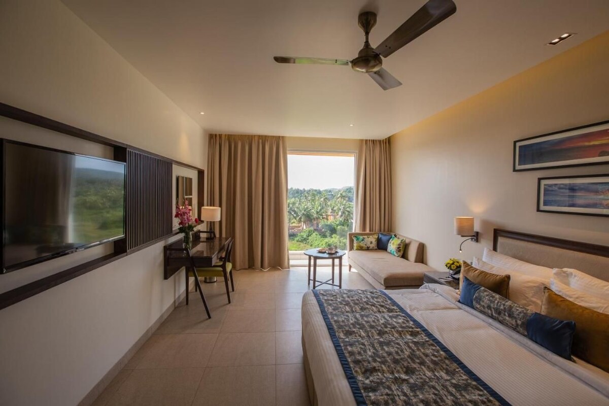 A hotel room with a bed, a desk, a chair, a TV, a ceiling fan, and a window with curtains.