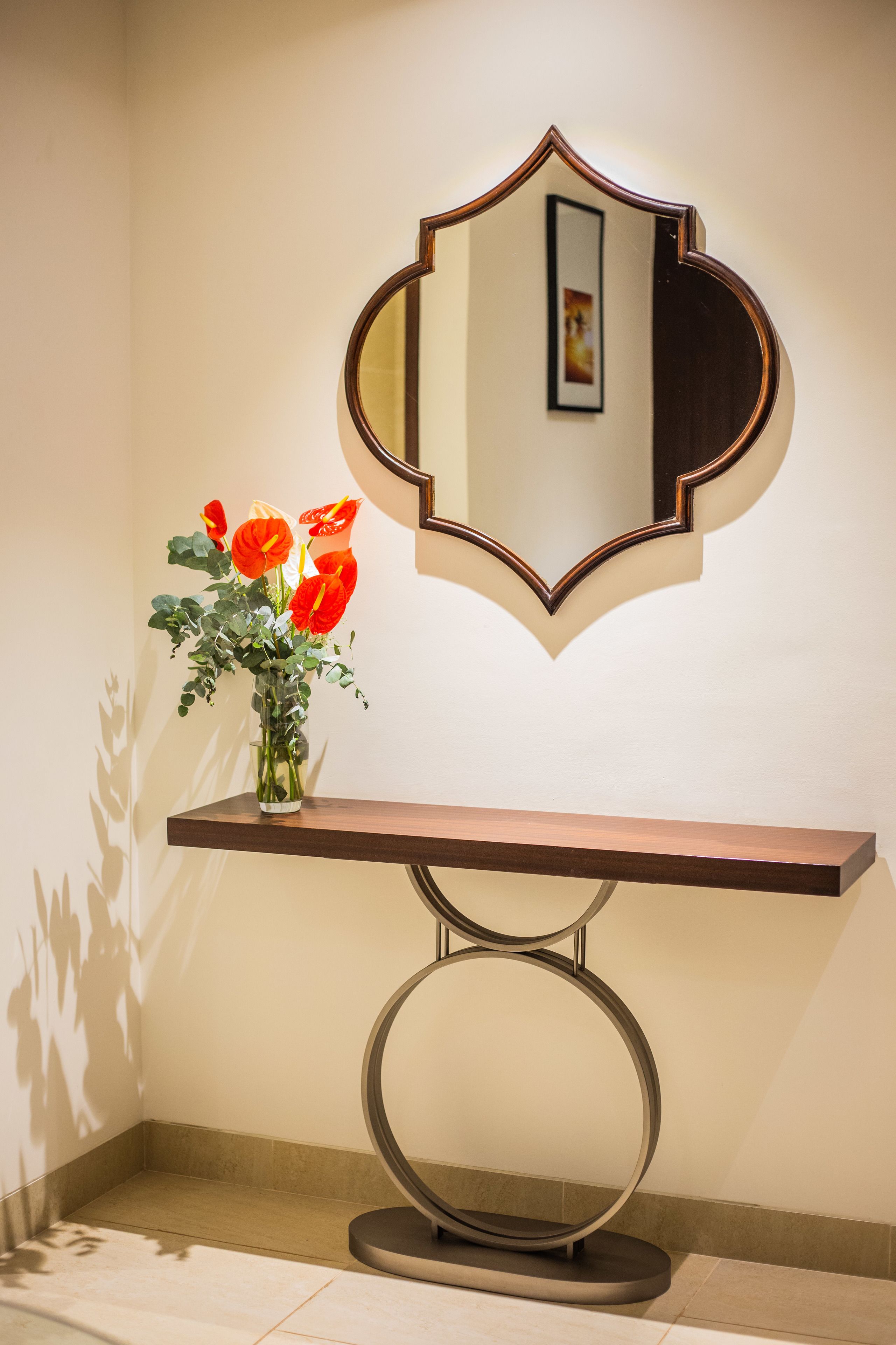 Un espejo de forma singular, una consola de madera y un florero con flores rojas.